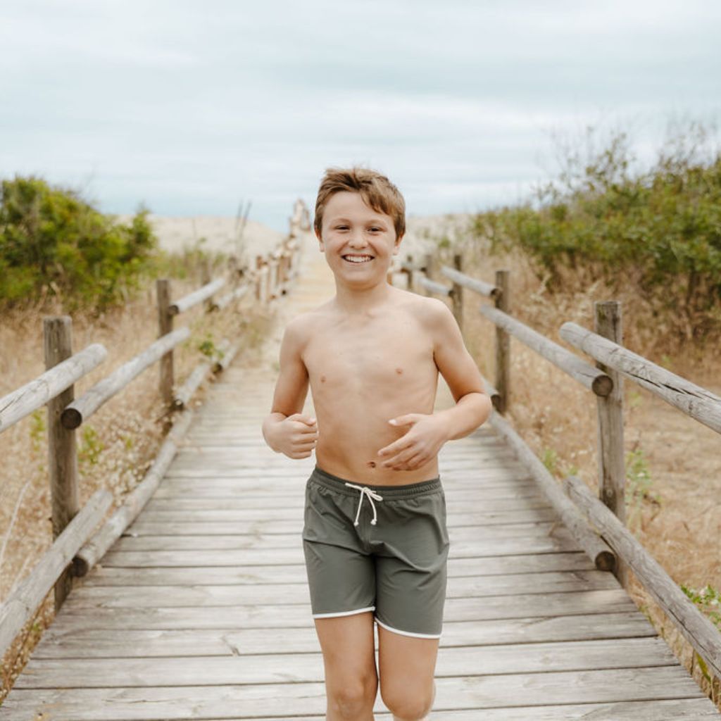 Little boy running along the boardwalk wearing the Folpetto Tommaso swim shorts in sage green and ivory