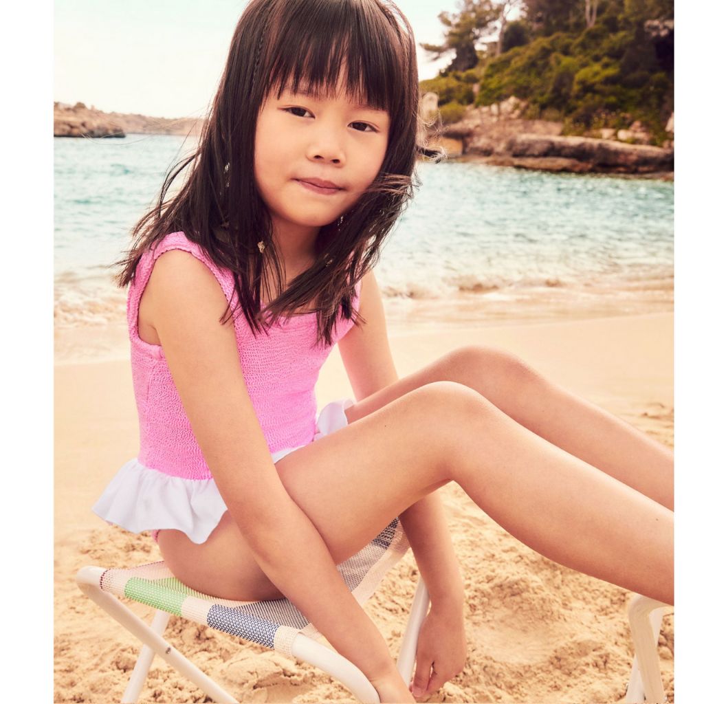 Little girl on the beach wearing the Hunza G Baby Denise swimsuit in bubblegum featuring the original crinkle