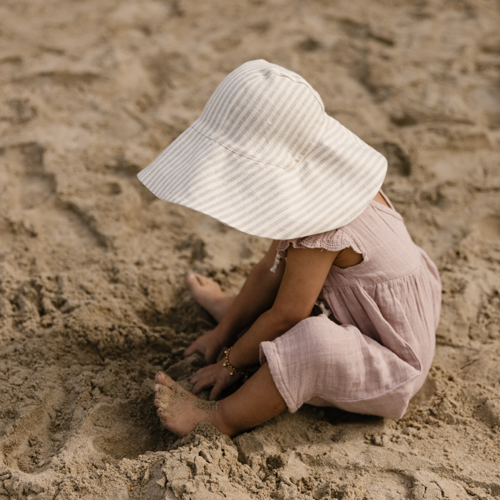 Little girl on the beach showing a side view of the Briar Baby sun hat in beige and grey Harbor Stripe