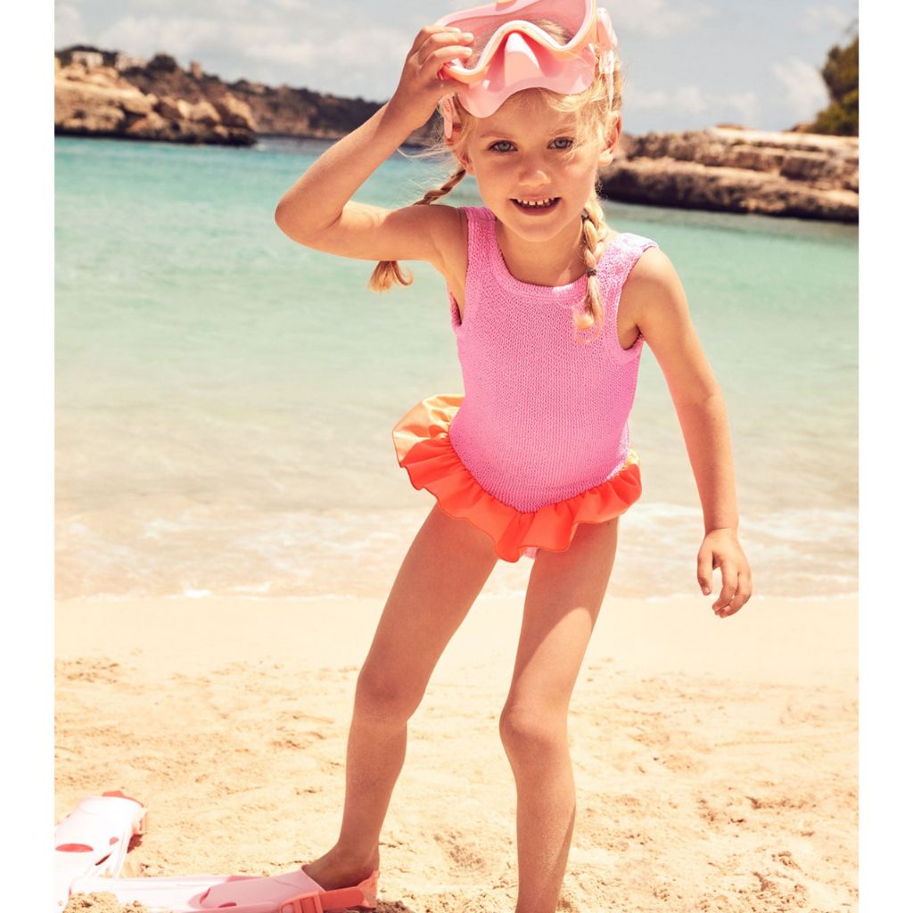 Little girl on the beach wearing the Hunza G Baby Duo Denise swimsuit in bubblegum and orange featuring the original crinkle