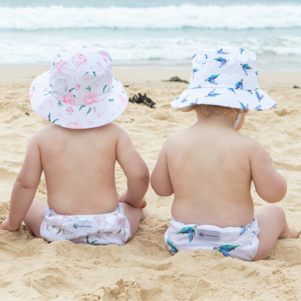 Children on the beach wearing Anchor & Arrow Sea Turtle and Pretty Peony print unisex reusable swim nappy and swim hat