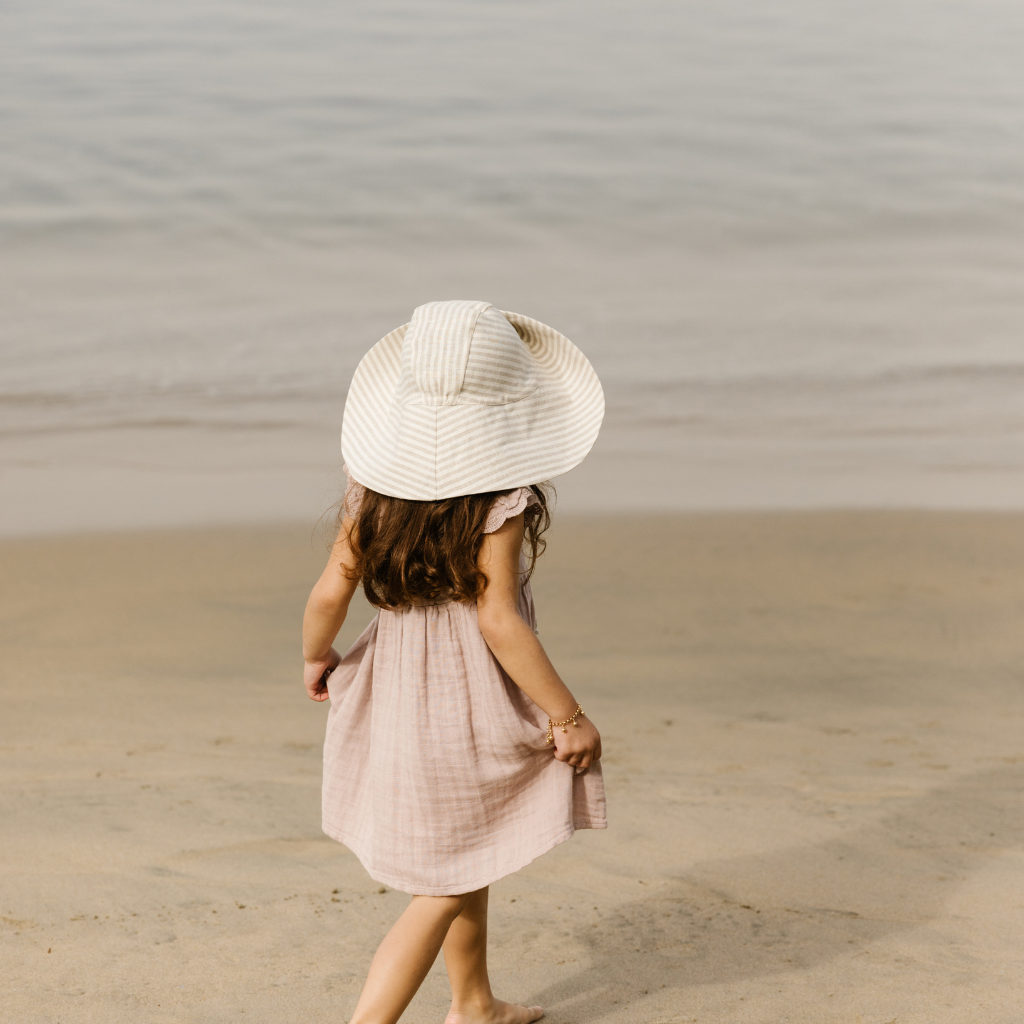 Little girl on the beach wearing a Briar Baby sun hat in beige and grey Harbor Stripe