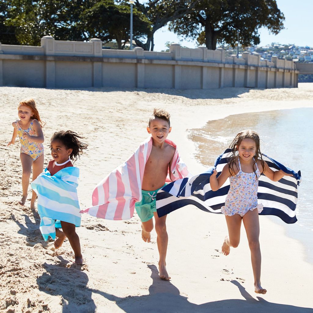 Children on the beach with their Dock and Bay Signature striped cabana towel in Malibu Pink, Tulum Blue and Whitsunday Blue