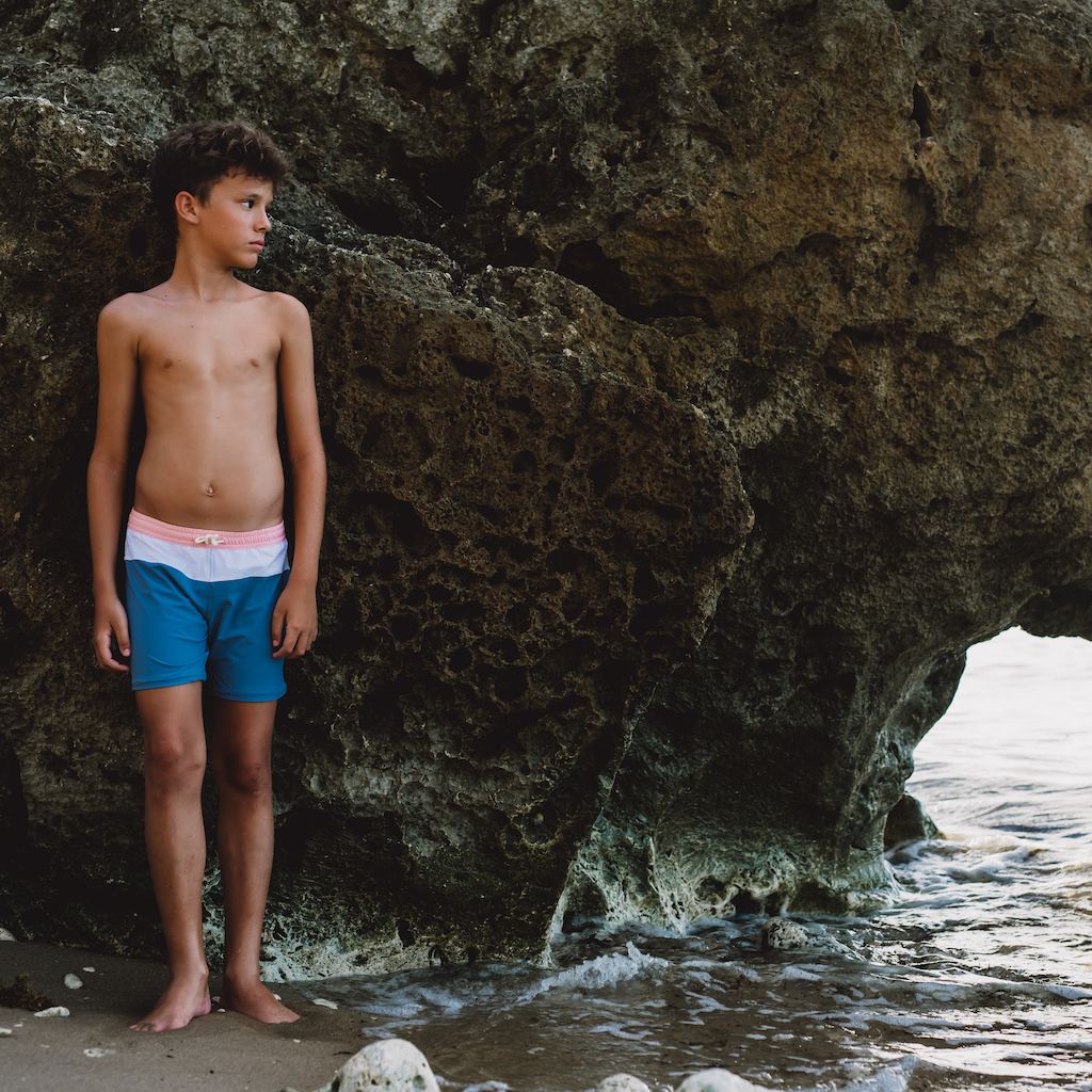 Boy by the sea wearing Folpetto Harry swim shorts for boys in blue and pink