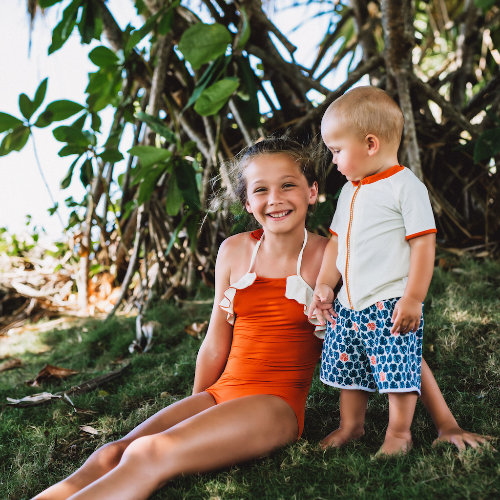 Baby boy wearing Folpetto Tommaso swim shorts for boys in teal blue and mandarin red tropical leaf print