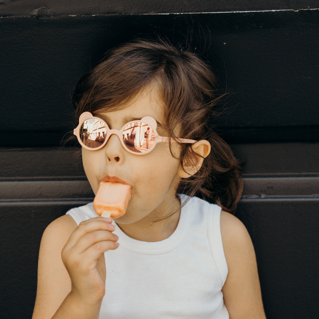 Little girl eating a lolly wearing Ki et La Ourson Teddy Bear sunglasses for children from 1 - 4 years in peach