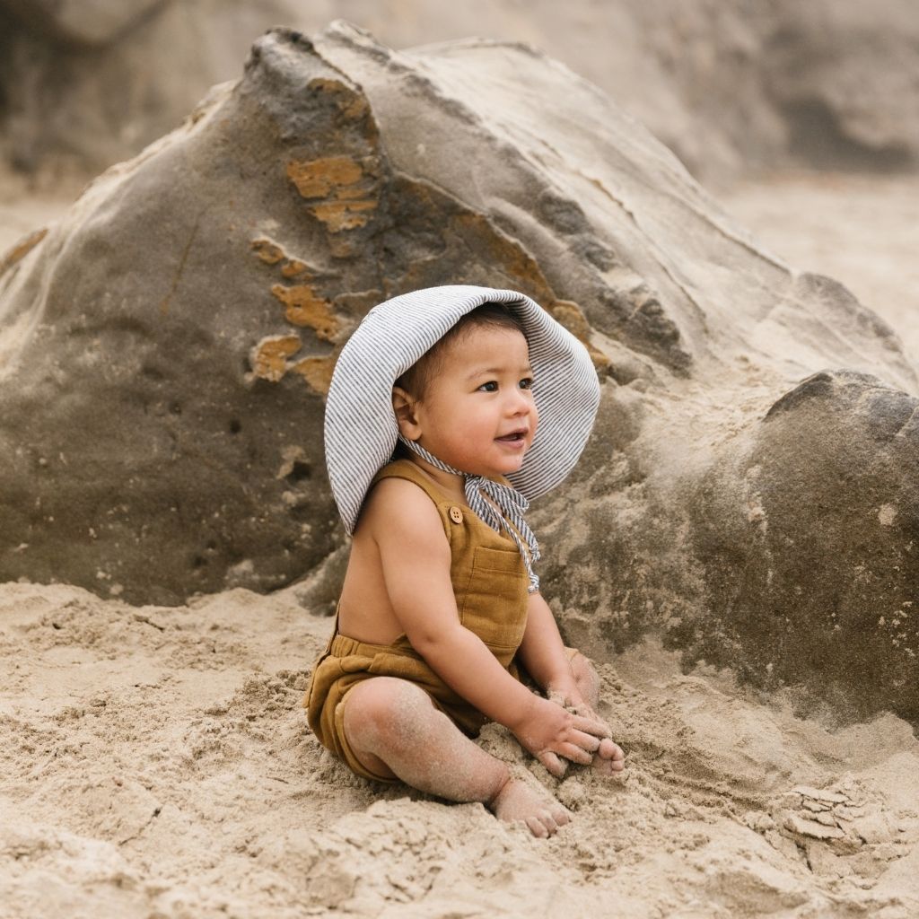 Little boy sitting on the beach wearing Unisex 100% linen baby sun bonnet from Briar Baby in navy and White Island Stripe