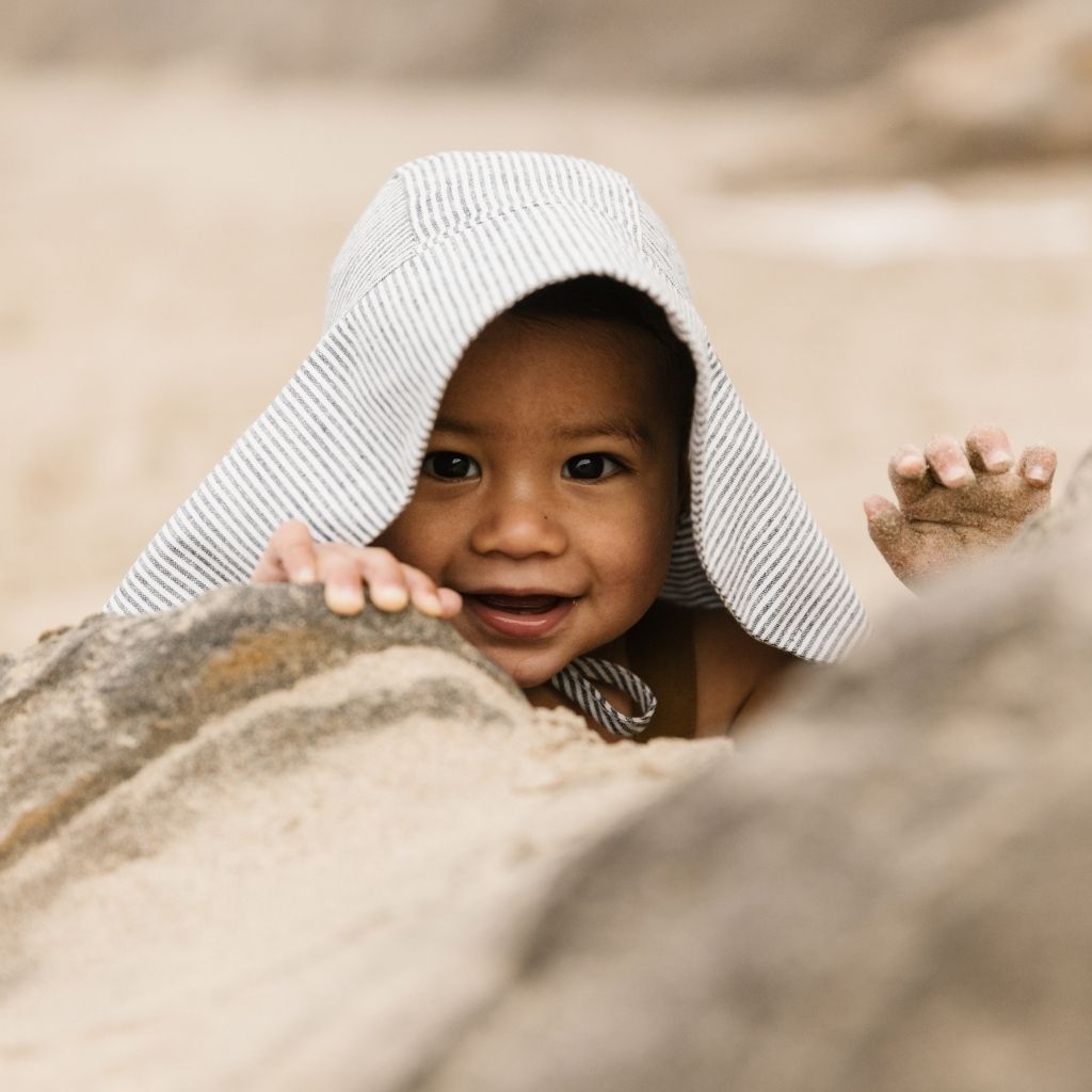 Little boy peeking through the rocks wearing Unisex 100% linen baby sun bonnet from Briar Baby in navy and White Island Stripe