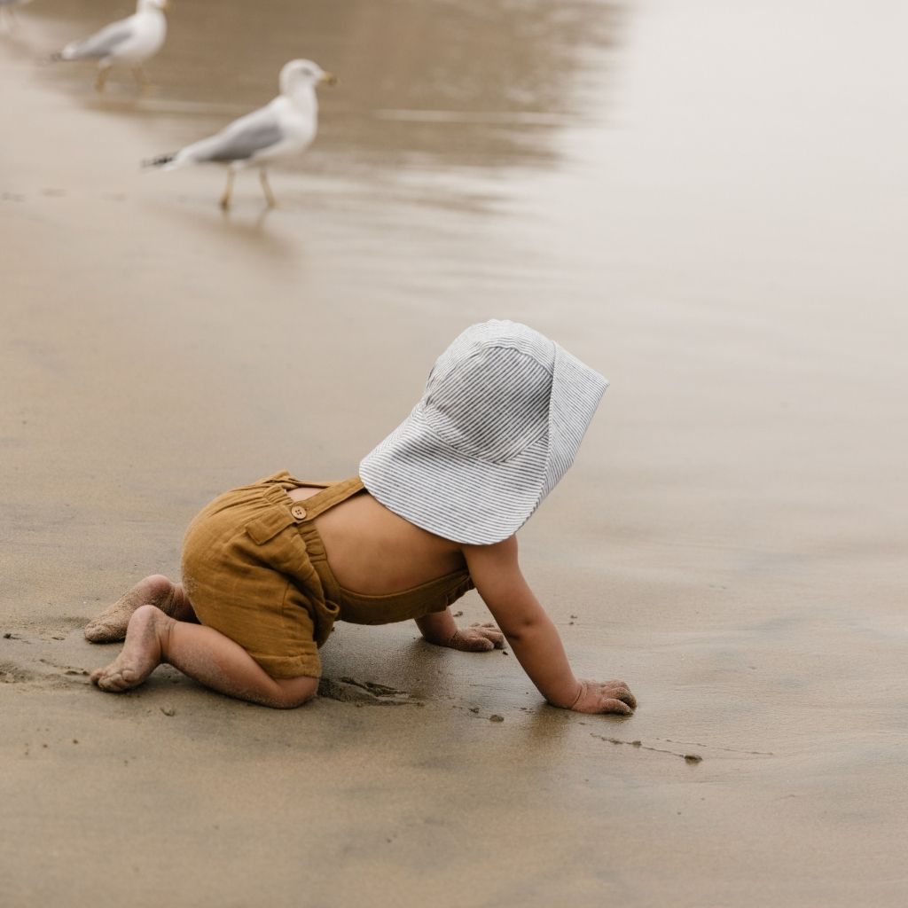 Little boy crawling in the wet sand wearing Unisex 100% linen baby sun bonnet from Briar Baby in navy and White Island Stripe