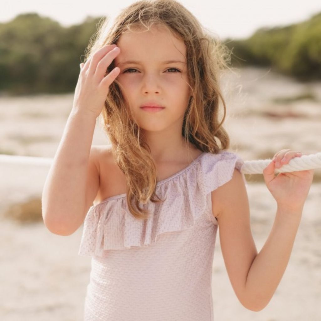 Little girl with her hand to her hair wearing the new Lison Paris Byblos One Shoulder Swimsuit in Pink Rose Gold