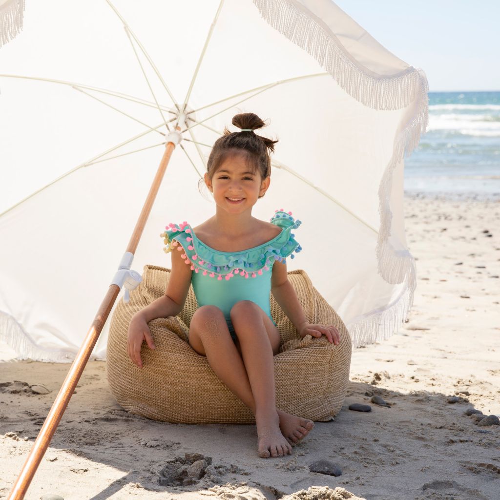 Little girl under a parasol on the beach wearing the PQ Swim Kids Seashore Pom Pom One Piece Ruffle Swimsuit