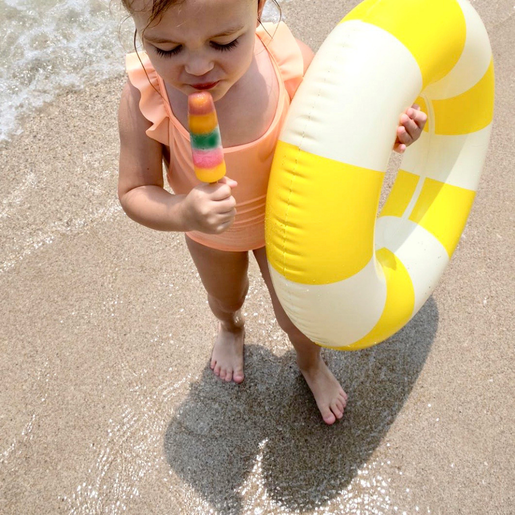 Little girl eating her lolly whilst holding the Petites Pommes Limonata yellow rubber ring inflatable