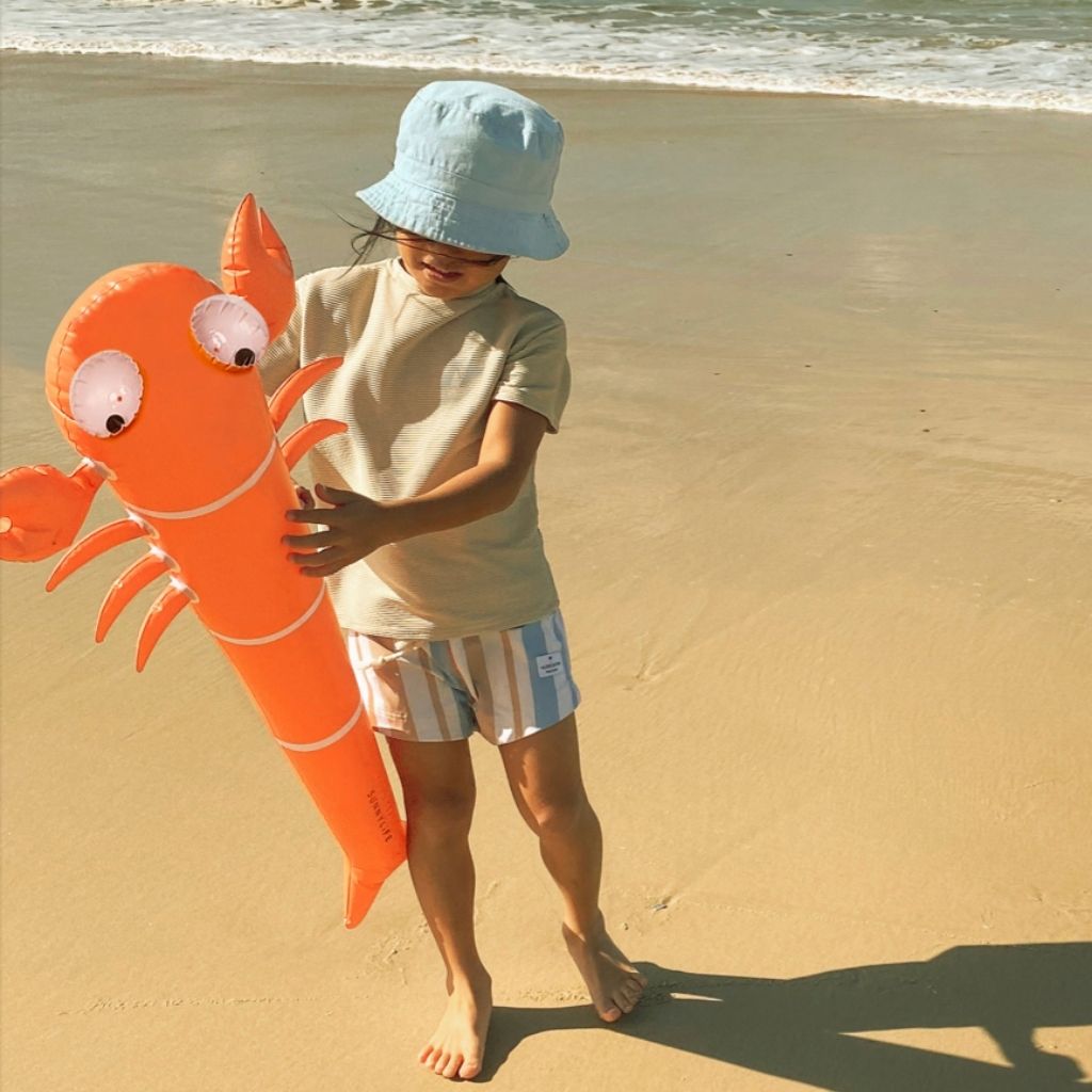 Little boy on the beach holding Sunnylife Kids inflatable noodle in sonny the sea creature neon orange set of two