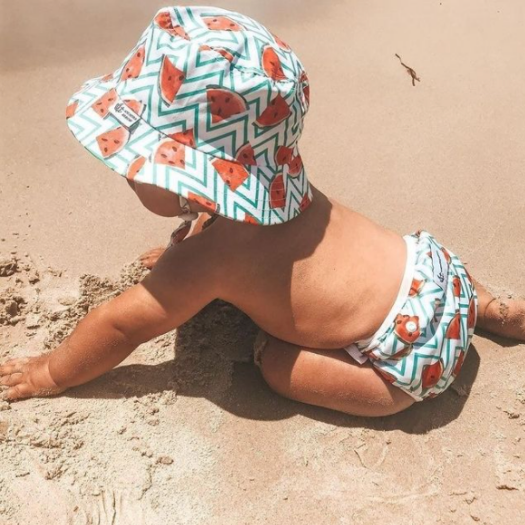 Baby in the sand wearing Anchor & Arrow Frolicking Watermelon print unisex reusable swim hat and swim nappy